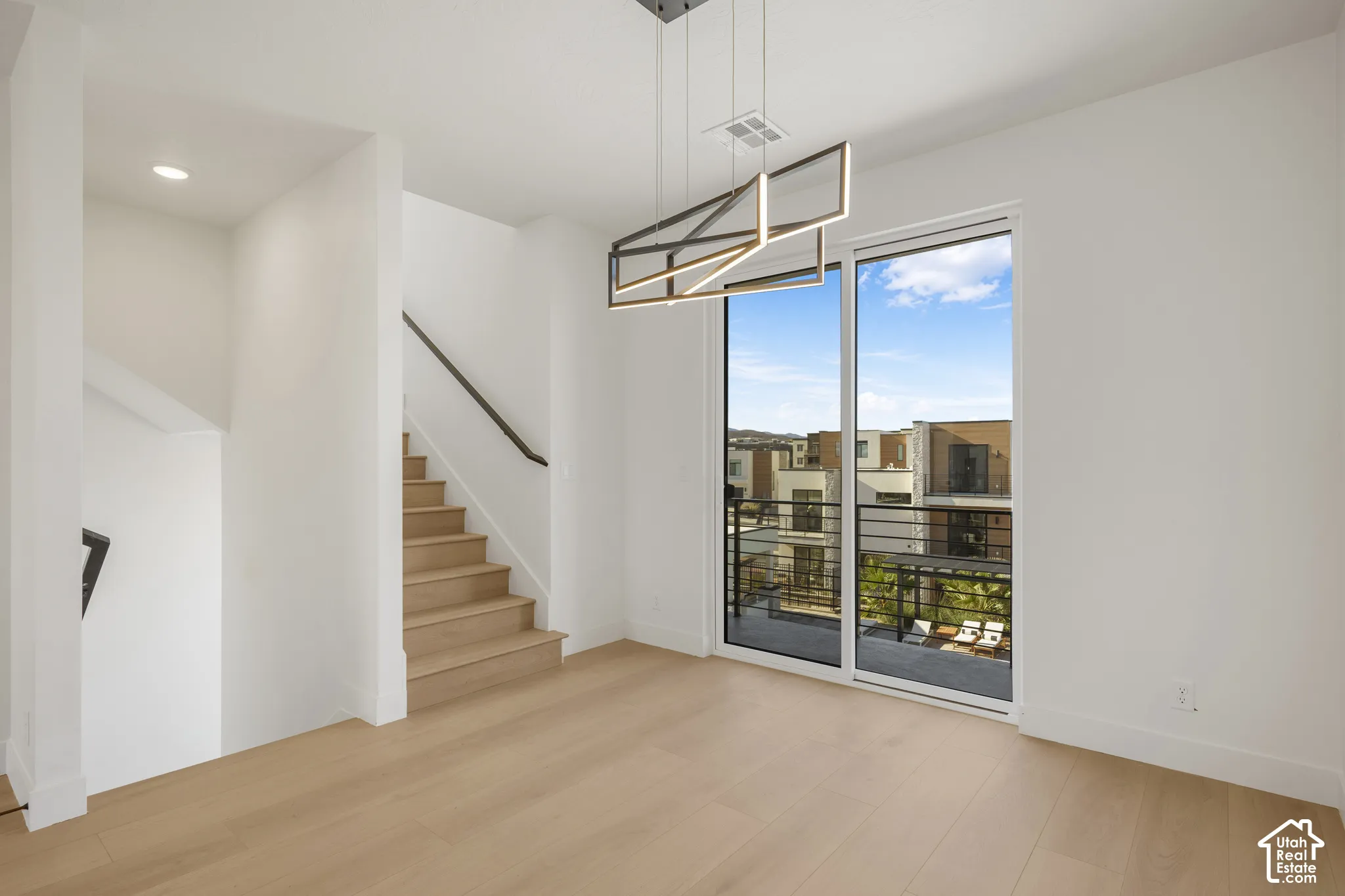 Open-concept interior featuring wood-finish flooring, a geometric linear pendant light, wood-tread staircase with a metal handrail, and floor-to-ceiling sliding glass doors leading to a balcony
