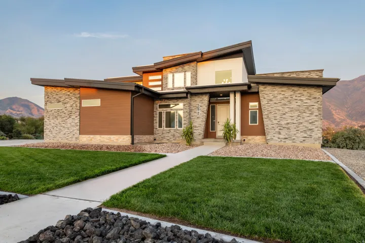Modern architectural exterior with a multi-level roofline, stone veneer and wood-finish siding, a concrete pathway, and a manicured lawn