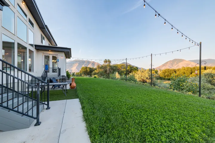 Expansive green lawn, string lighting, concrete patio, exterior staircase with metal railings, and a two-story residence with large windows