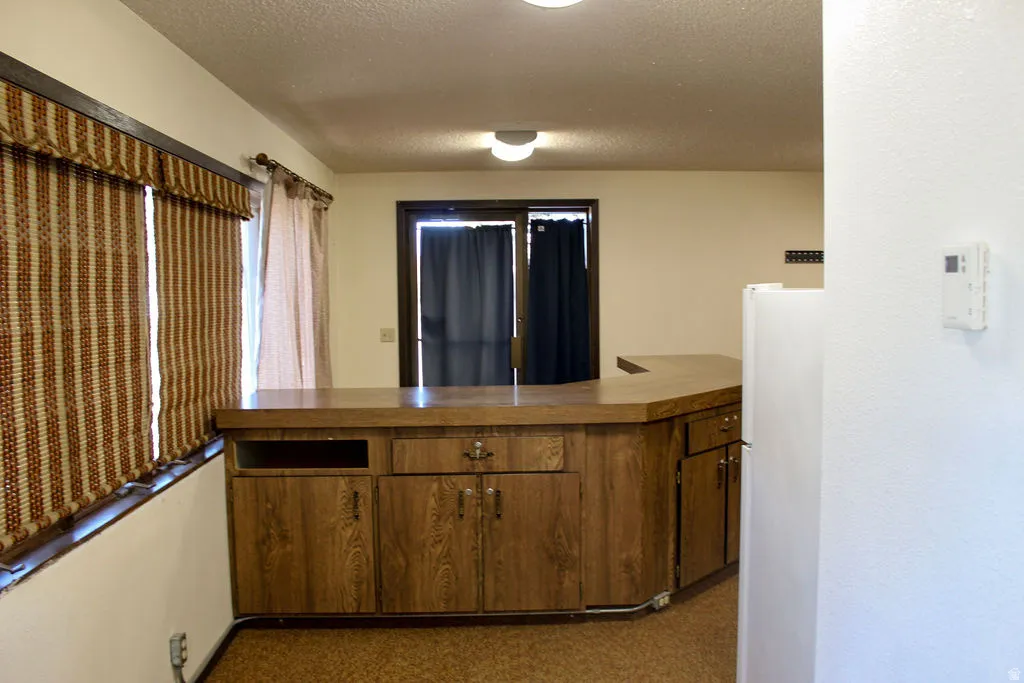 Kitchen featuring freestanding refrigerator, a textured ceiling, a peninsula, wood finish cabinetry, and dark colored carpet