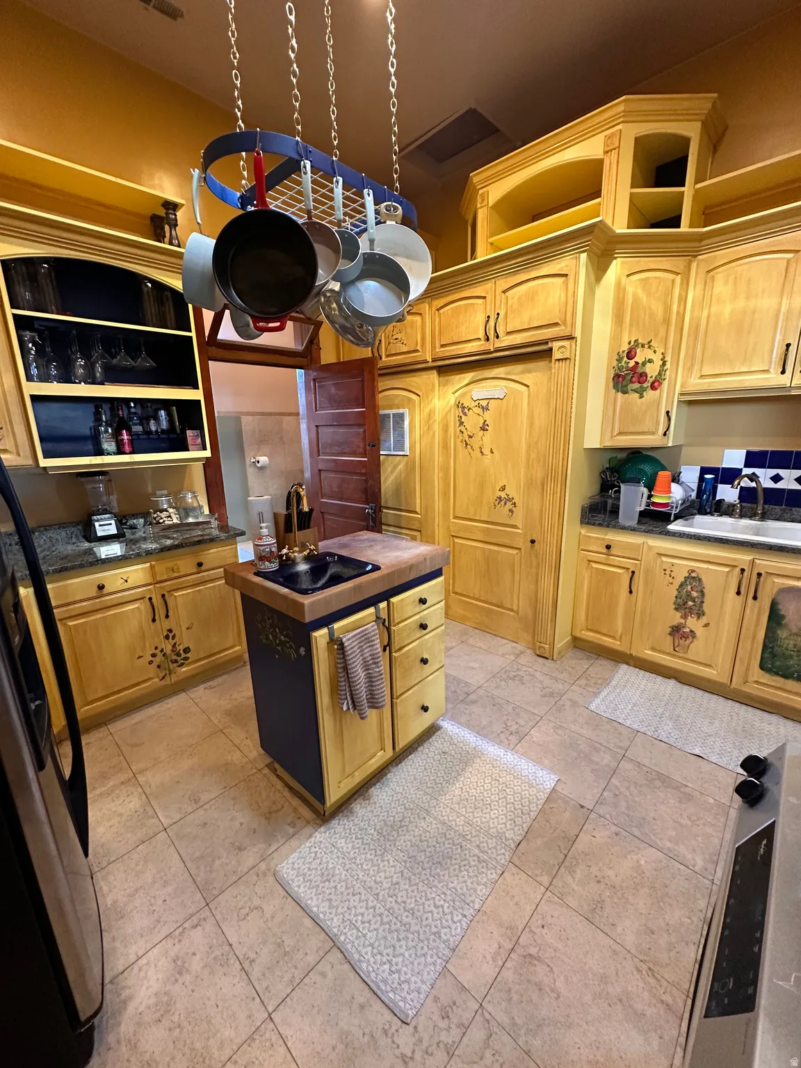 Kitchen with dark countertops, freestanding refrigerator, a kitchen island with sink, and light wood finish cabinetry