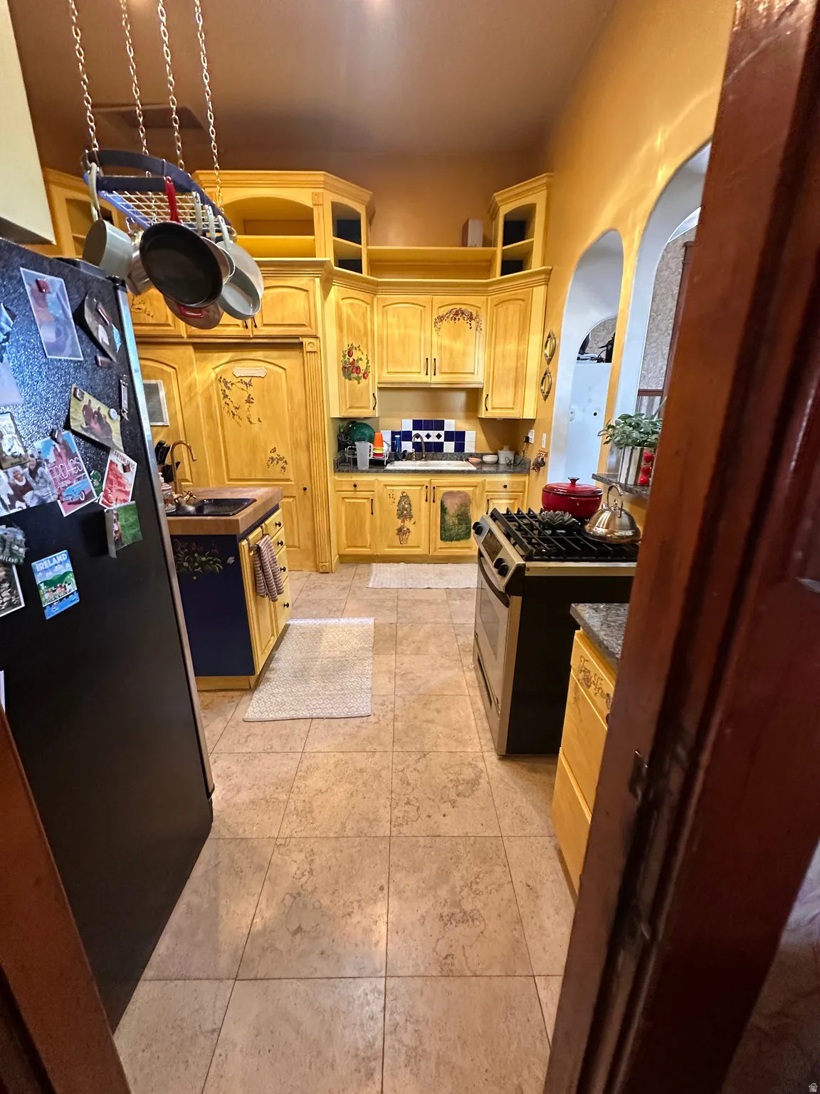 Kitchen with light wood finish cabinetry, freestanding refrigerator, stainless steel range with gas stovetop, dark countertops, and arched walkways