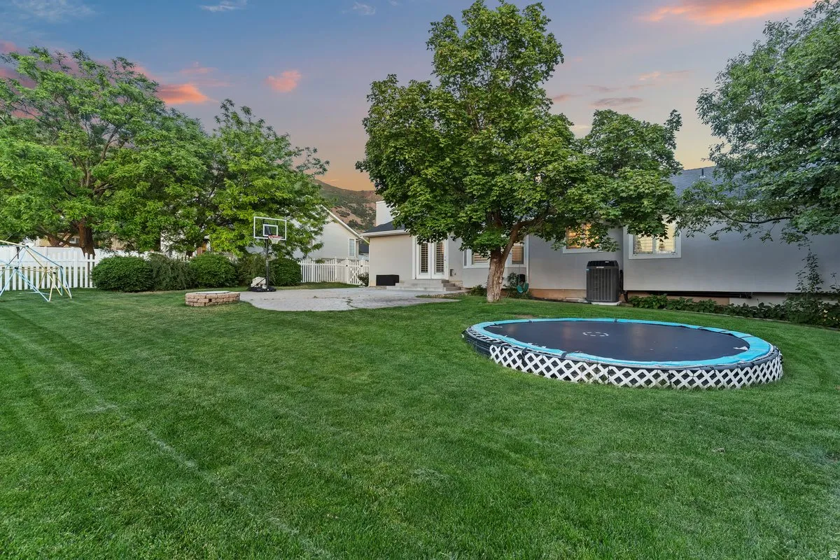 Expansive backyard featuring a large green lawn, mature trees, a concrete patio, and a white picket fence