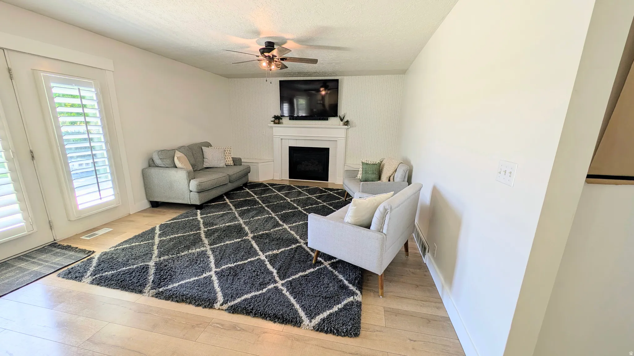 Spacious living area featuring light wood-finish flooring, a white mantel fireplace, and a ceiling fan with integrated lighting