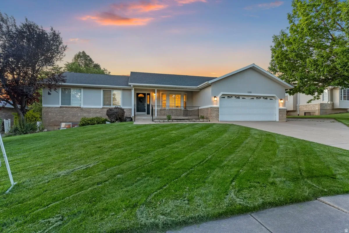 Ranch-style residence featuring a gray exterior with brick accents, an attached two-car garage, a covered front porch with railing, a concrete driveway, and a sloped front lawn