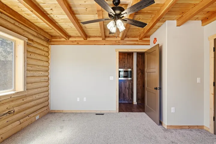 Unfurnished bedroom with log walls, carpet floors, a wooden ceiling with exposed beams, and ceiling fan