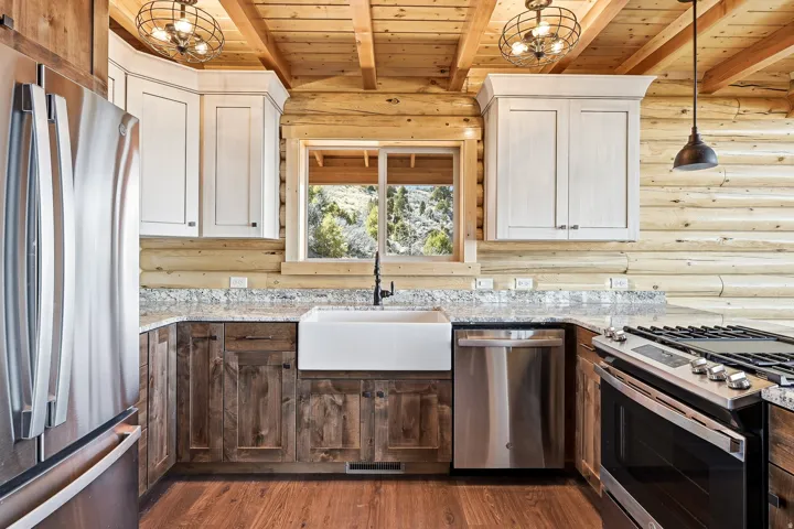 Two tone kitchen featuring suspended lighting, stainless steel appliances, light stone counters, rustic walls, and a wood ceiling with exposed beams