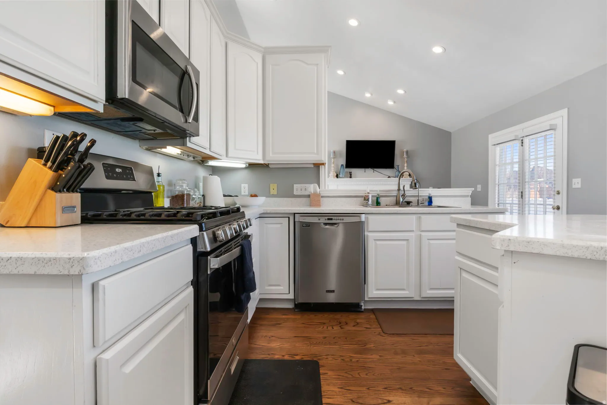Kitchen featuring stainless steel appliances, lofted ceiling, white cabinets, light stone counters, and dark wood-type flooring