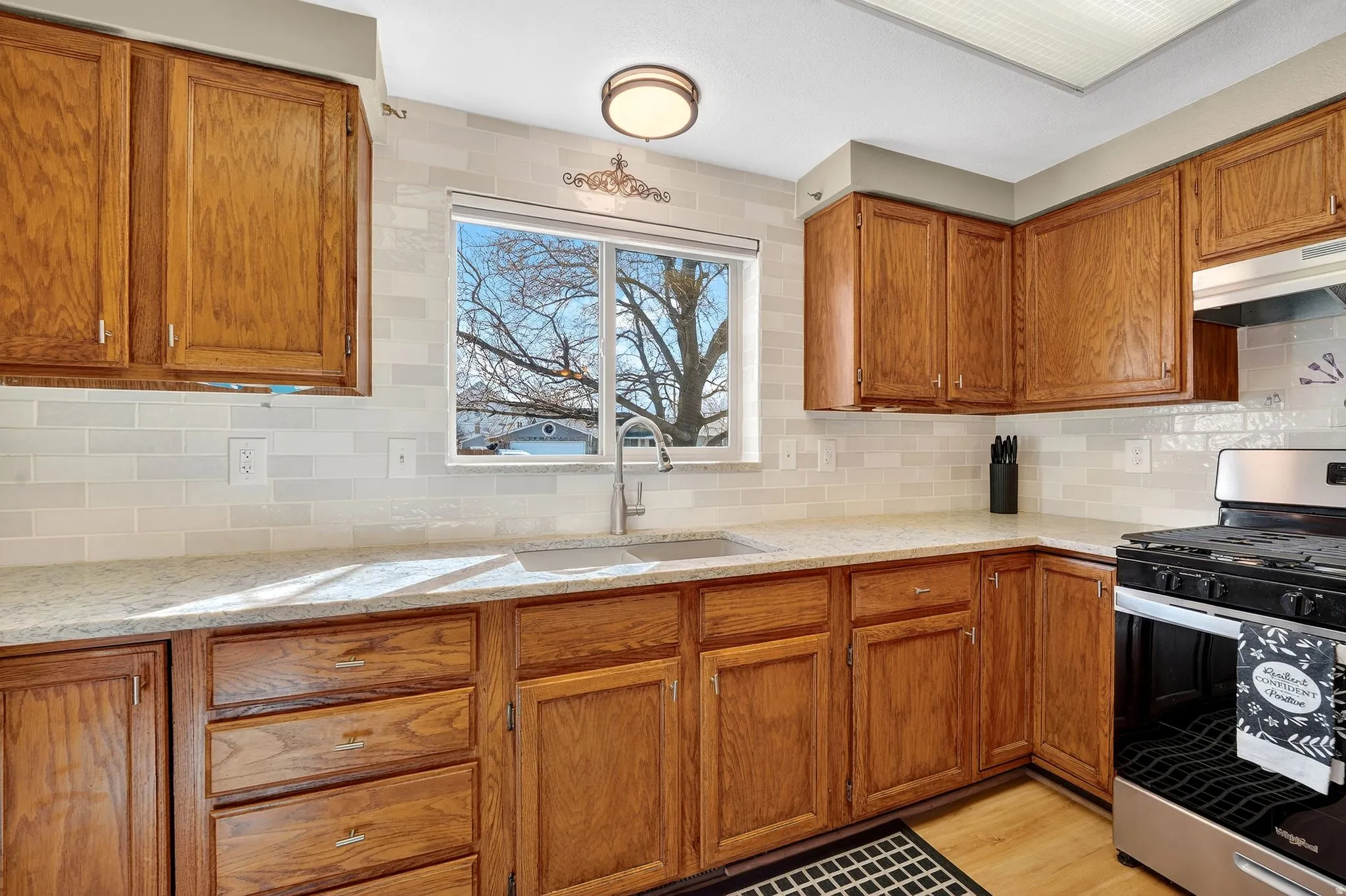 Kitchen with stainless steel gas stove, wood finish cabinetry, light stone countertops, decorative backsplash, and light wood-style floors