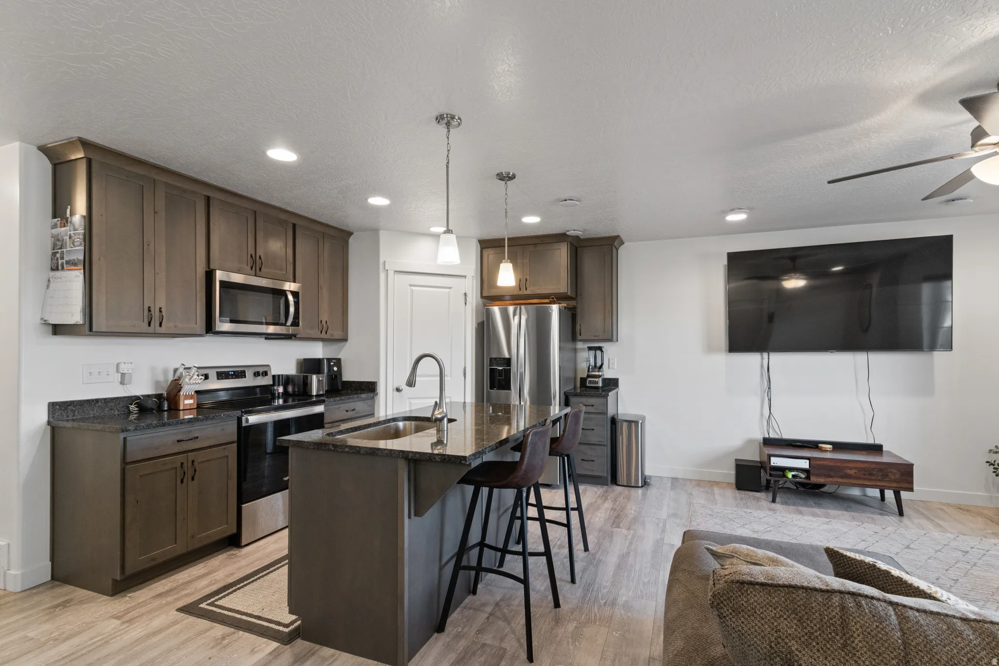 Kitchen featuring stainless steel appliances, dark stone counters, dark wood finish cabinetry, an island with sink, and a textured ceiling