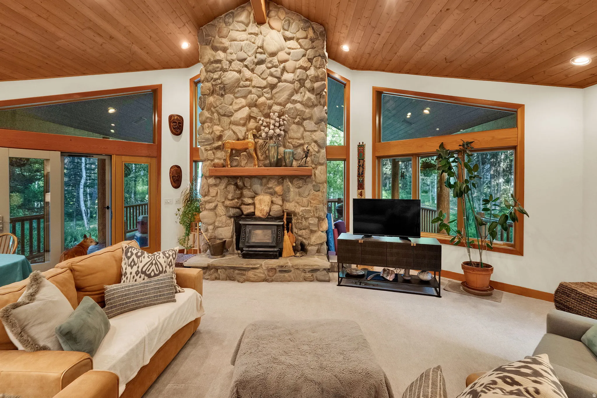 Stone fireplace with wood-burning stove, vaulted wood-paneled ceiling, large windows with wood frames, and glass-paned doors leading to an exterior space