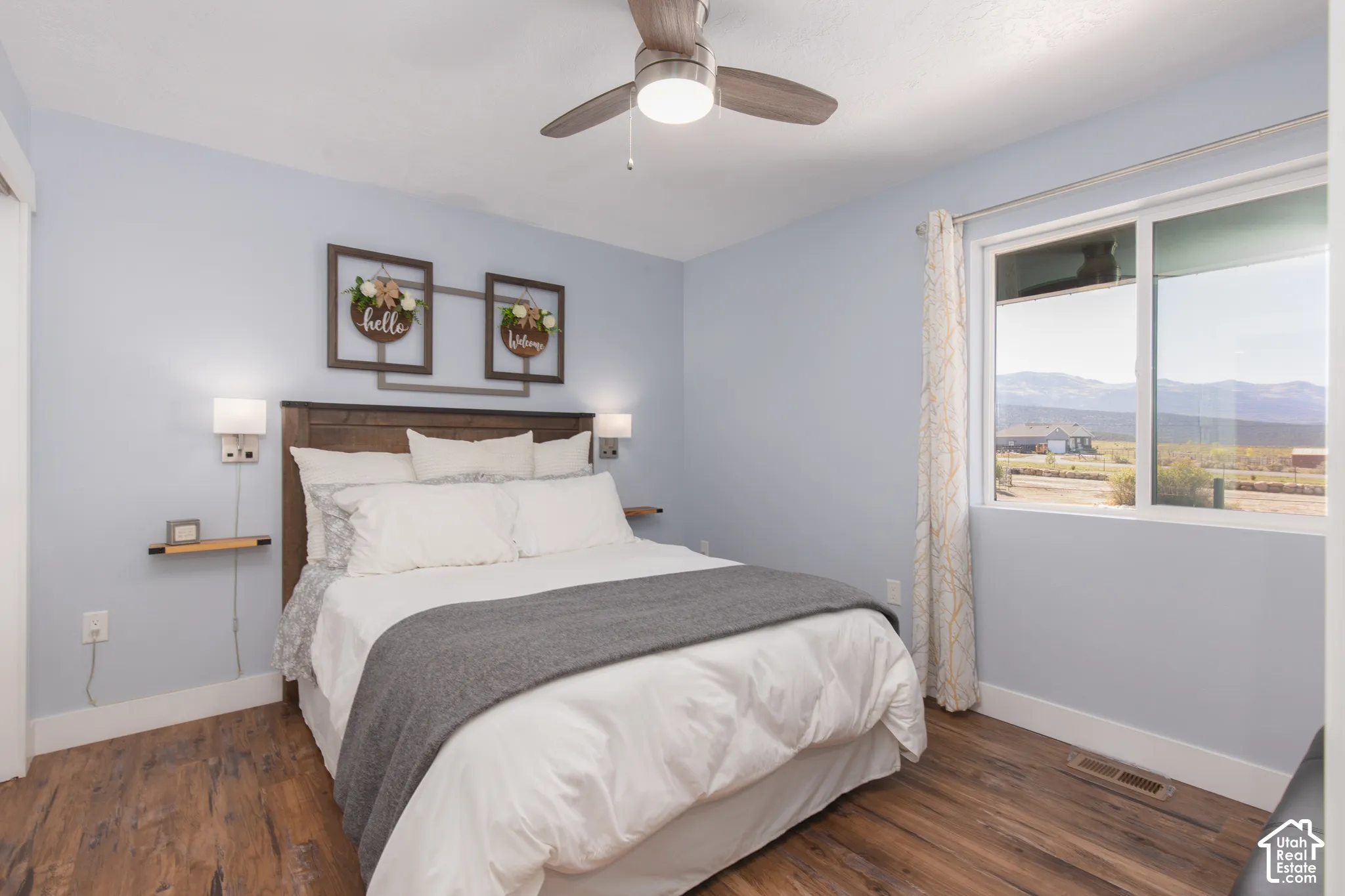 Wood-finish flooring, ceiling fan with integrated lighting, window with mountain scenery, and a wood headboard with integrated sconces and floating shelves