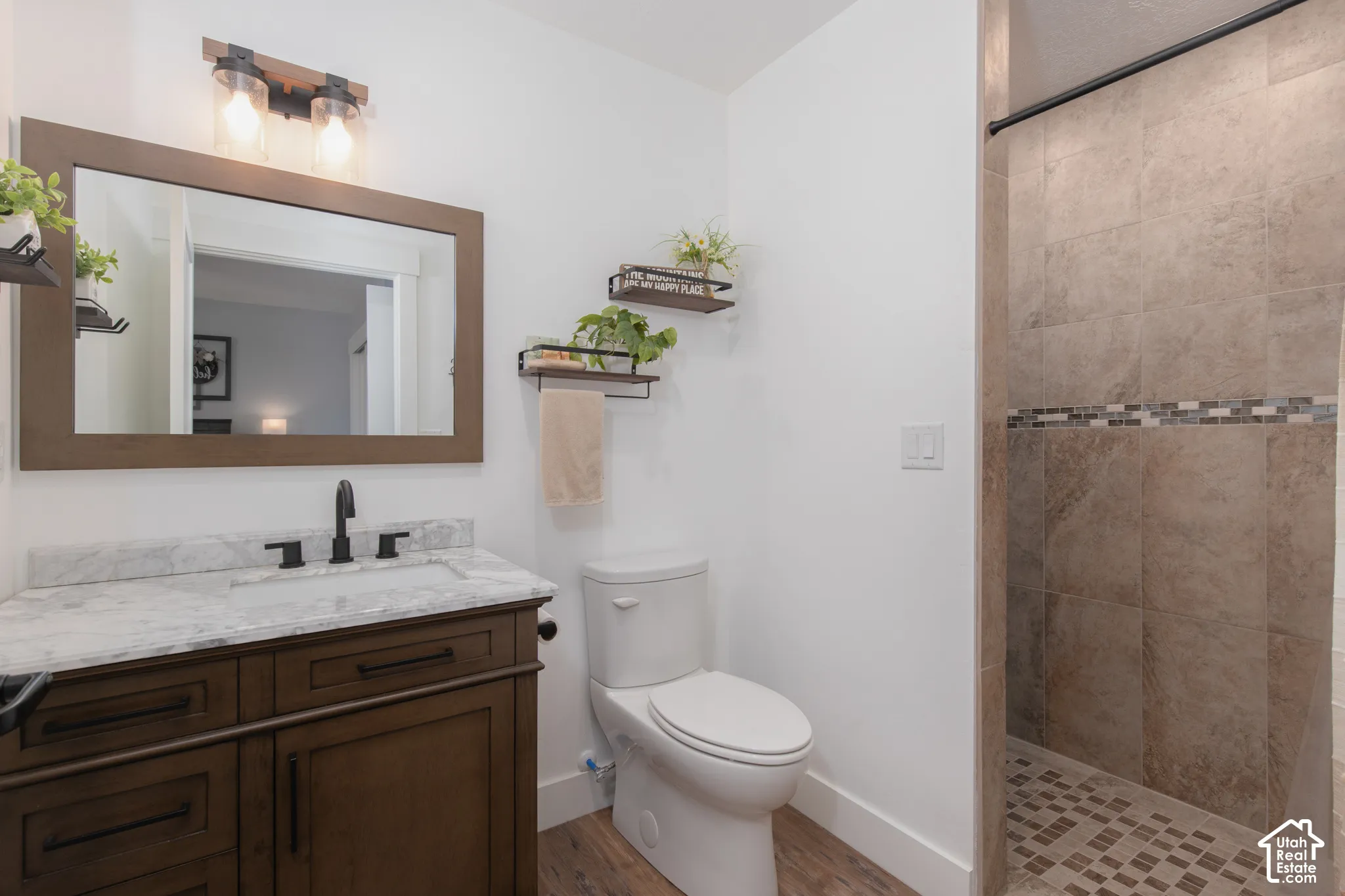 Wood-finish flooring, dark wood vanity with a white countertop, rectangular undermount sink with a matte black faucet, and a tiled walk-in shower with mosaic accents