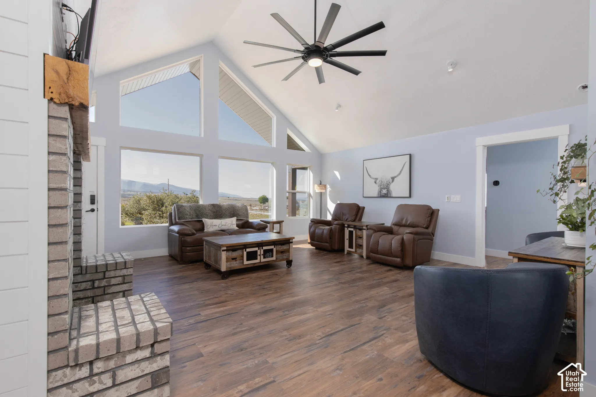 Vaulted ceiling with a contemporary ceiling fan, expansive windows, light blue walls, wood-finish flooring, and a brick fireplace with a wood mantel
