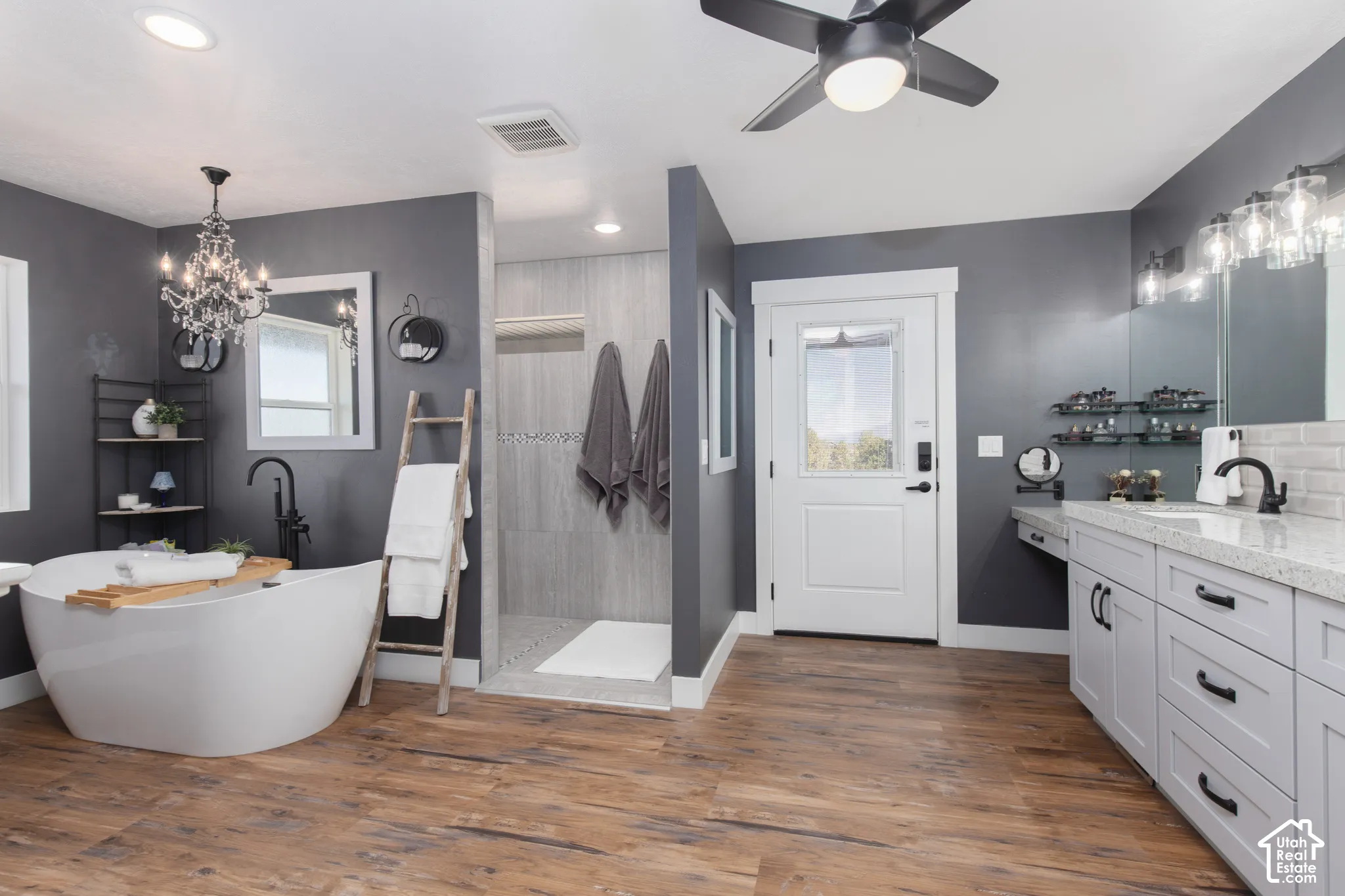 Freestanding soaking tub, walk-in shower with tiled walls, wood-finish flooring, dual vanity with light-colored countertops, and a ceiling fan