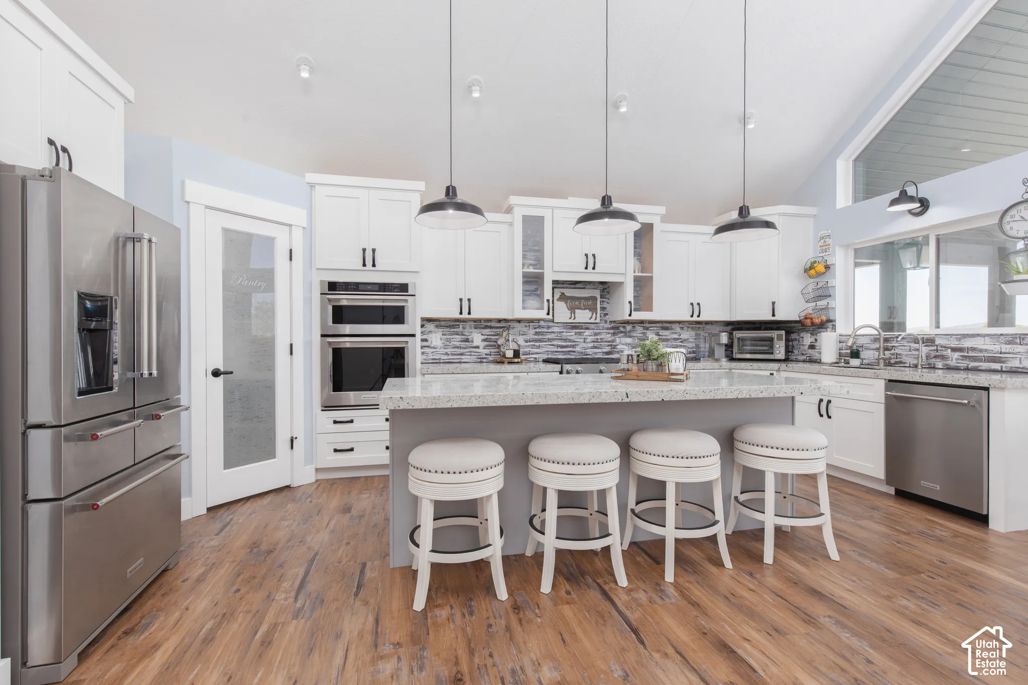 Wood-finish flooring, white cabinetry, stainless steel appliances, central island with seating, and a vaulted ceiling with accent window