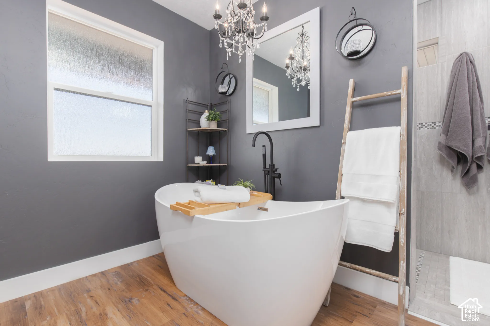 Freestanding soaking tub with a matte black faucet, wood-finish flooring, a white-framed window, and a chandelier fixture