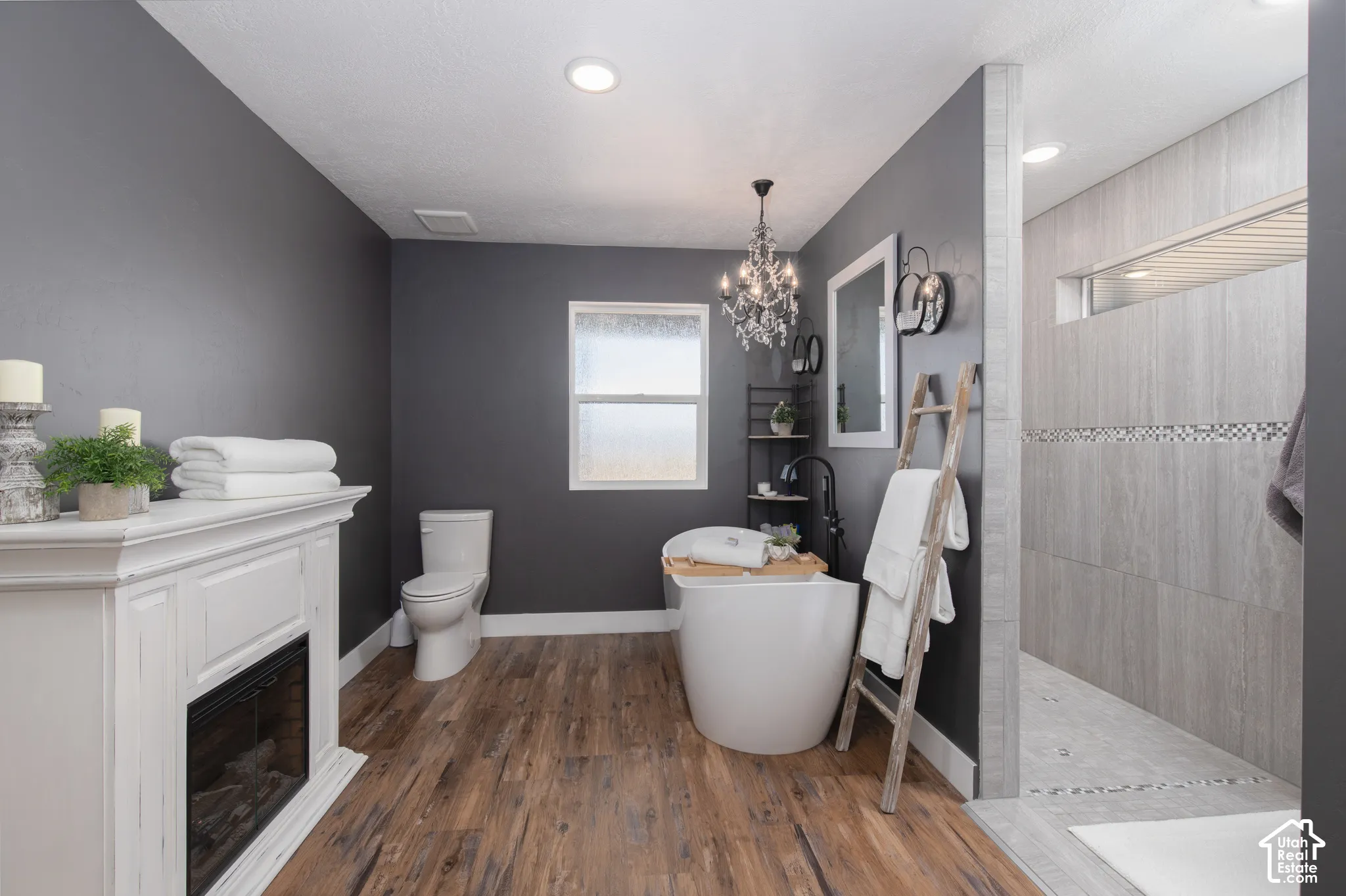 Wood-finish flooring, freestanding bathtub with dark-finish faucet, white built-in fireplace, tile-lined shower, and a chandelier