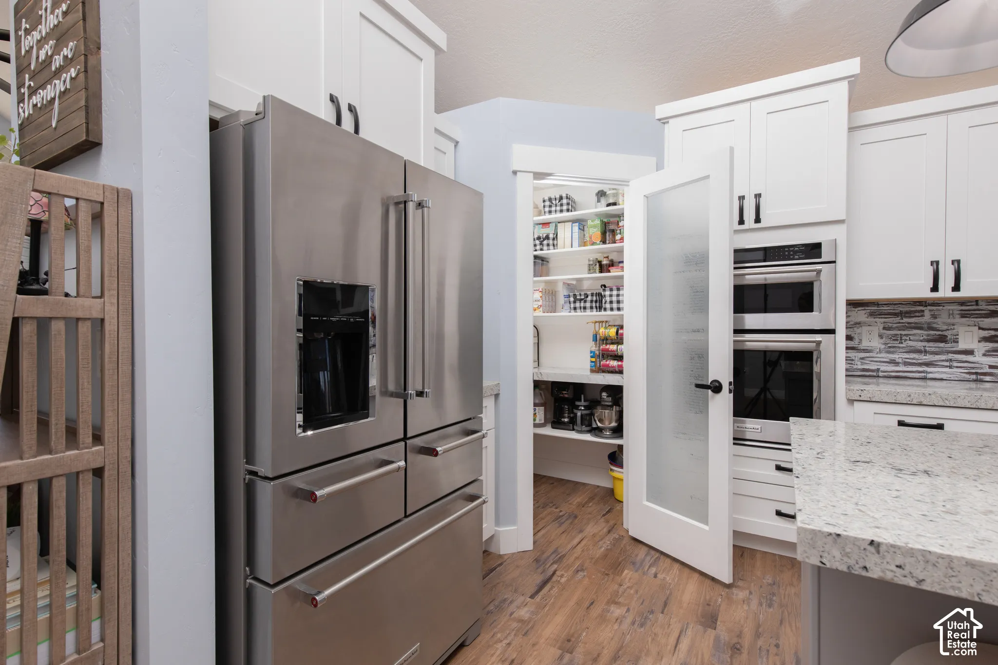 Stainless steel refrigerator, built-in oven and microwave, white shaker-style cabinetry with matte black hardware, light-toned countertop, and wood-finish flooring