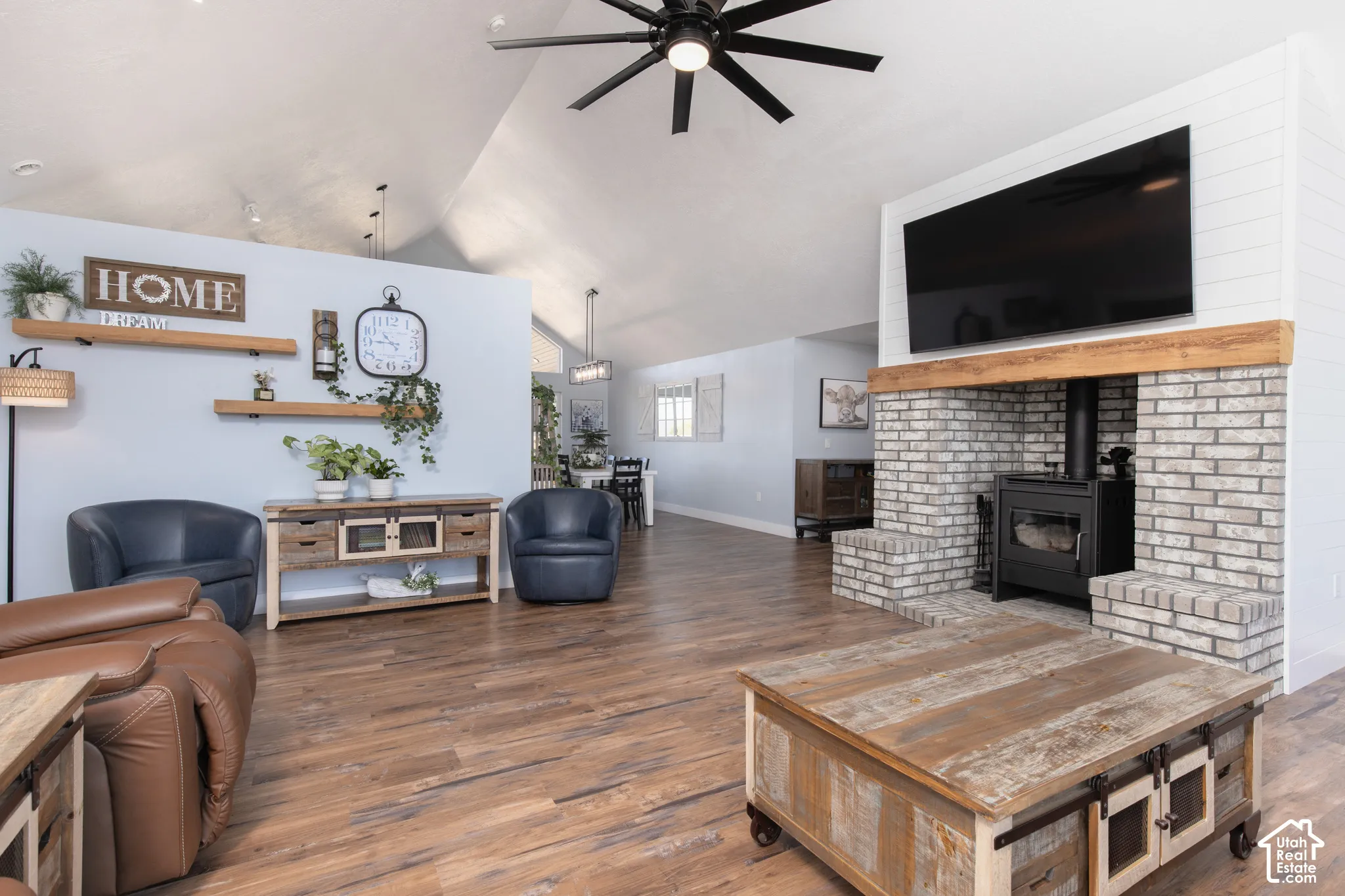 Vaulted ceiling, wood-finish flooring, brick-faced wood-burning stove, exposed wood mantel, and black ceiling fan