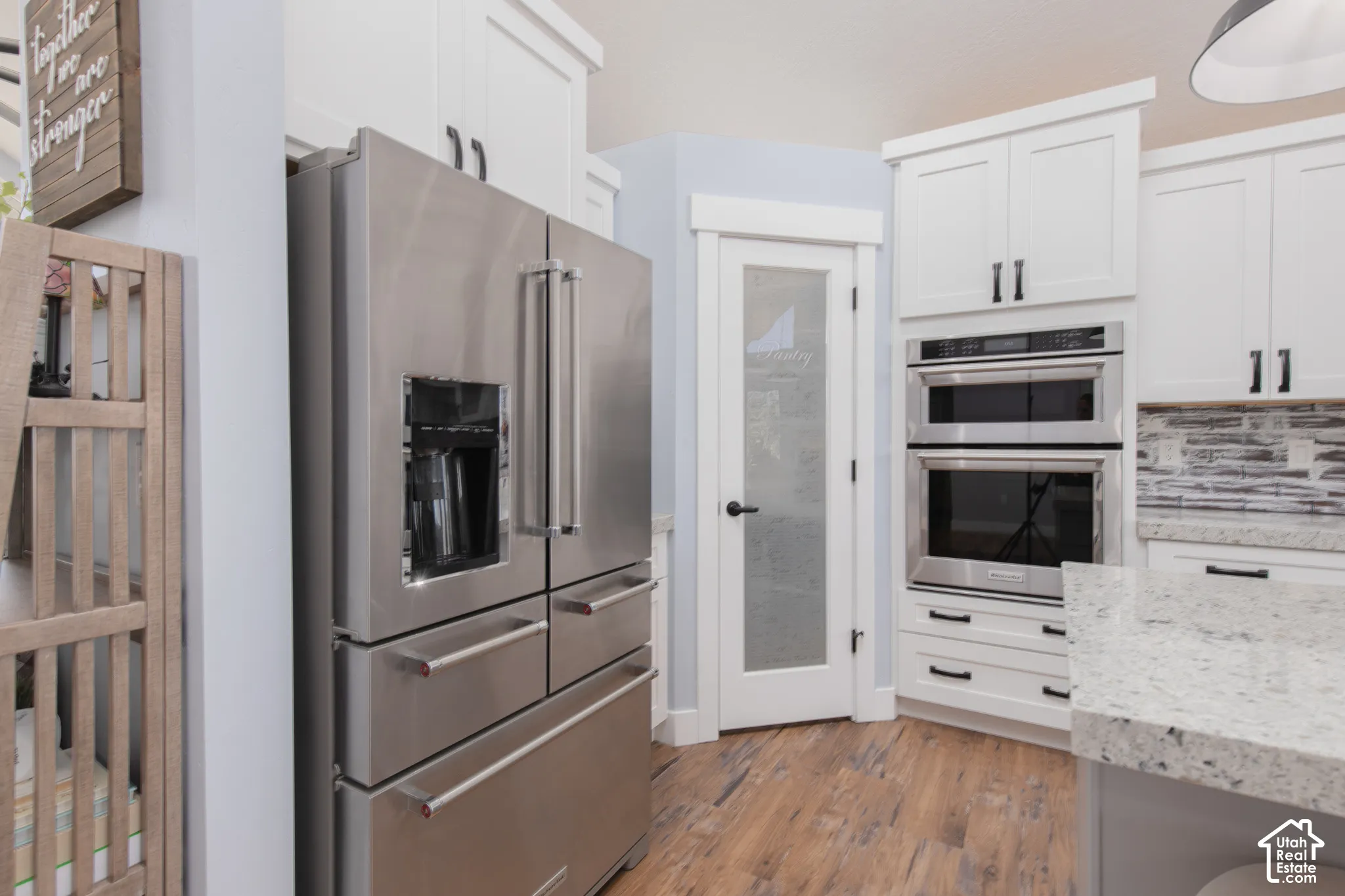 Stainless steel French door refrigerator, white shaker-style cabinetry with matte black hardware, built-in double ovens, frosted glass pantry door, and wood-finish flooring