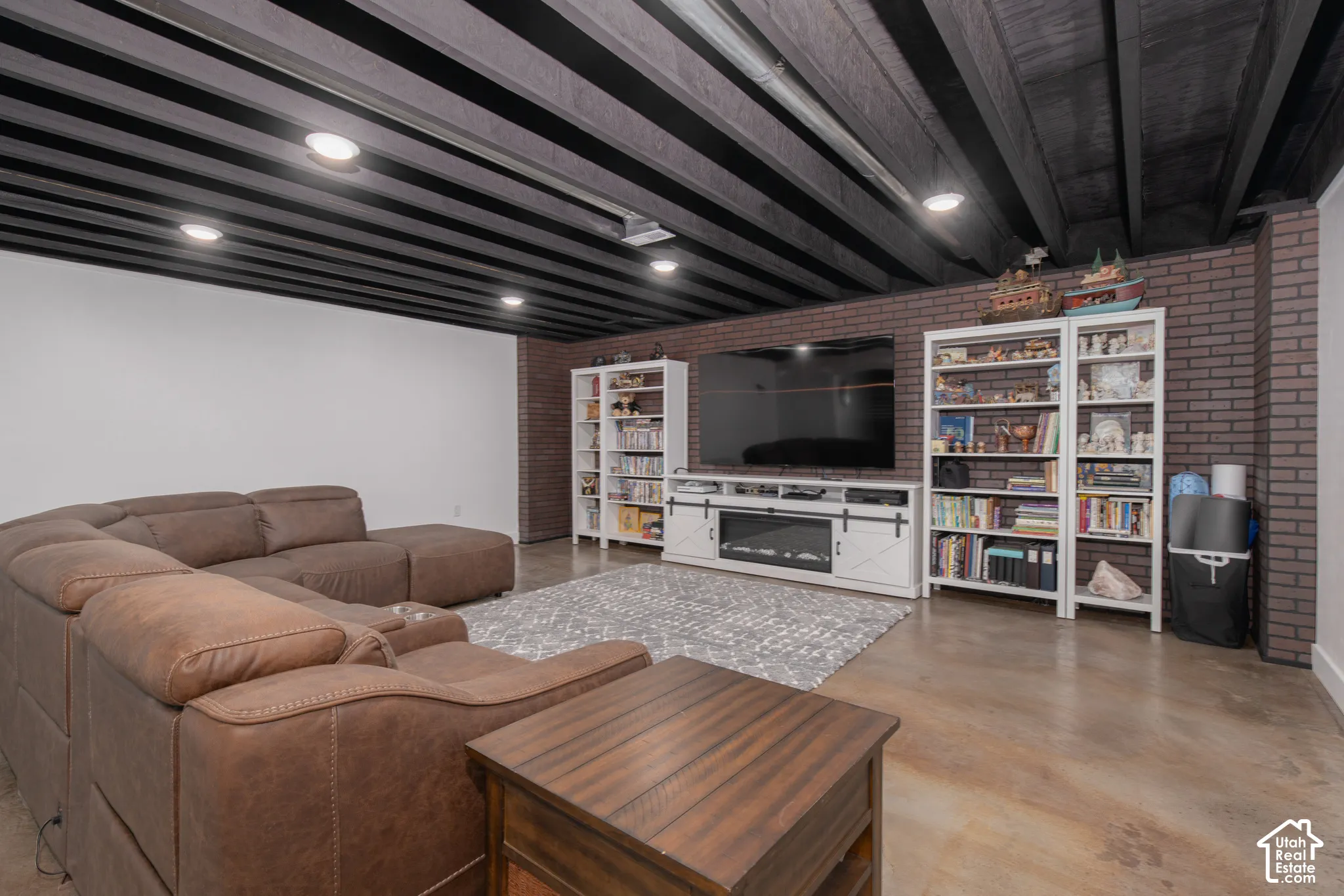 Exposed ceiling joists, recessed lighting, brick accent wall, built-in entertainment center, and concrete-finish flooring