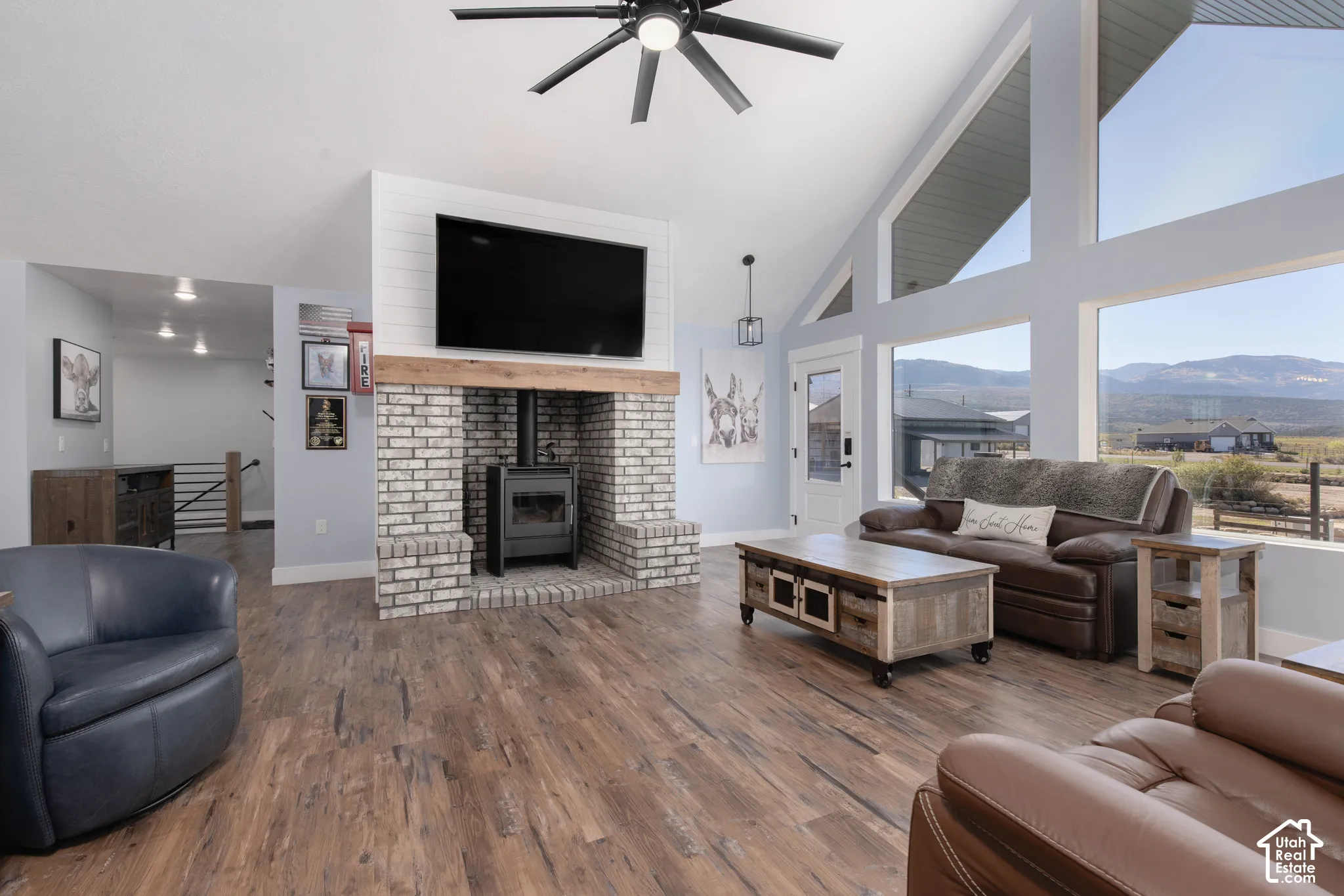 Vaulted ceilings, wood-finish flooring, white brick fireplace with wood mantel, large picture windows, and ceiling fan