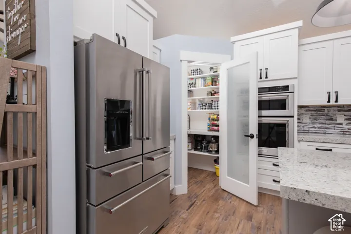 Stainless steel refrigerator, built-in oven and microwave, white shaker-style cabinetry with matte black hardware, light-toned countertop, and wood-finish flooring