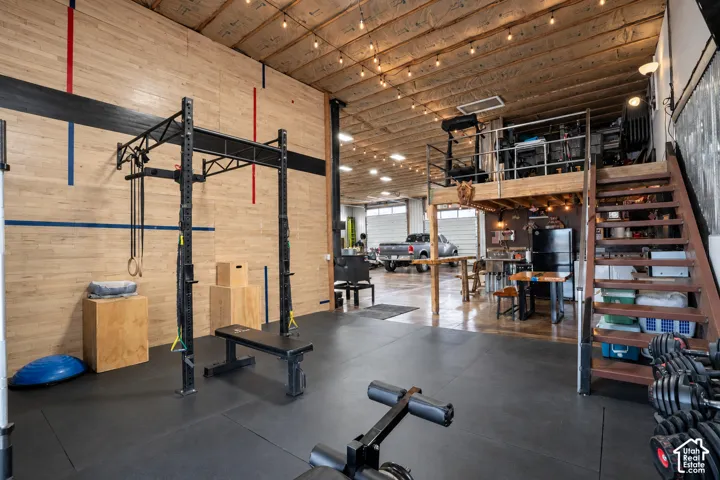 High ceilings with exposed wood beams, industrial-style string lighting, a dedicated fitness area with rubber flooring, a built-in mezzanine, and a functional staircase with integrated storage