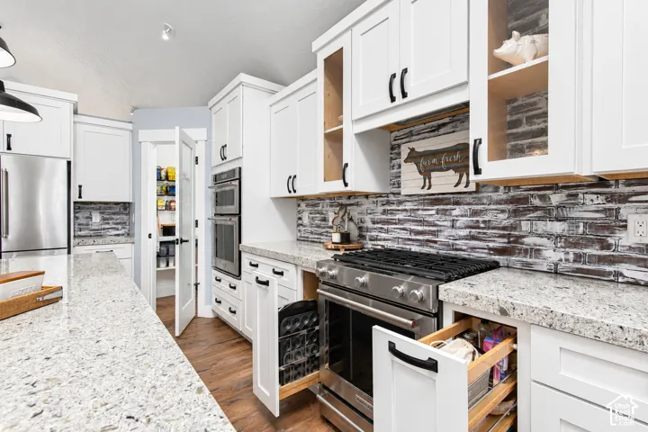White shaker cabinetry, stone countertops, stainless steel appliances, distressed brick-patterned backsplash, and wood-finish flooring