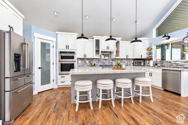Kitchen featuring wood-finish flooring, white cabinetry with black hardware, stainless steel appliances, a light-colored backsplash, and a central island with a contrasting base