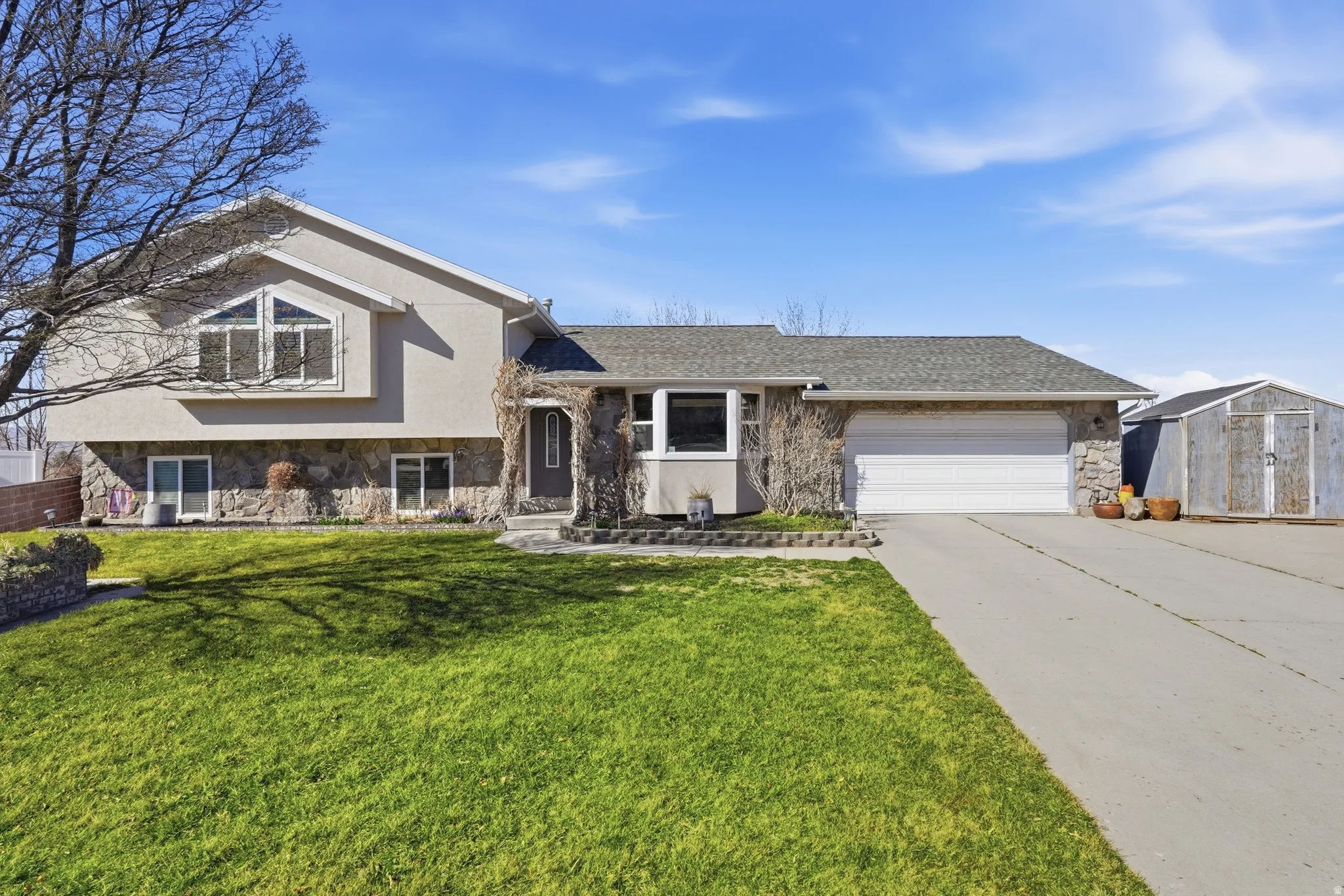 Split level home featuring stone siding, driveway, an attached garage, a front yard, and stucco siding