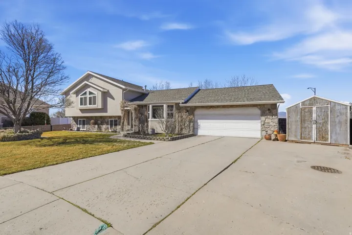 Split level home featuring stone siding, an attached garage, concrete driveway, a front lawn, and a storage unit