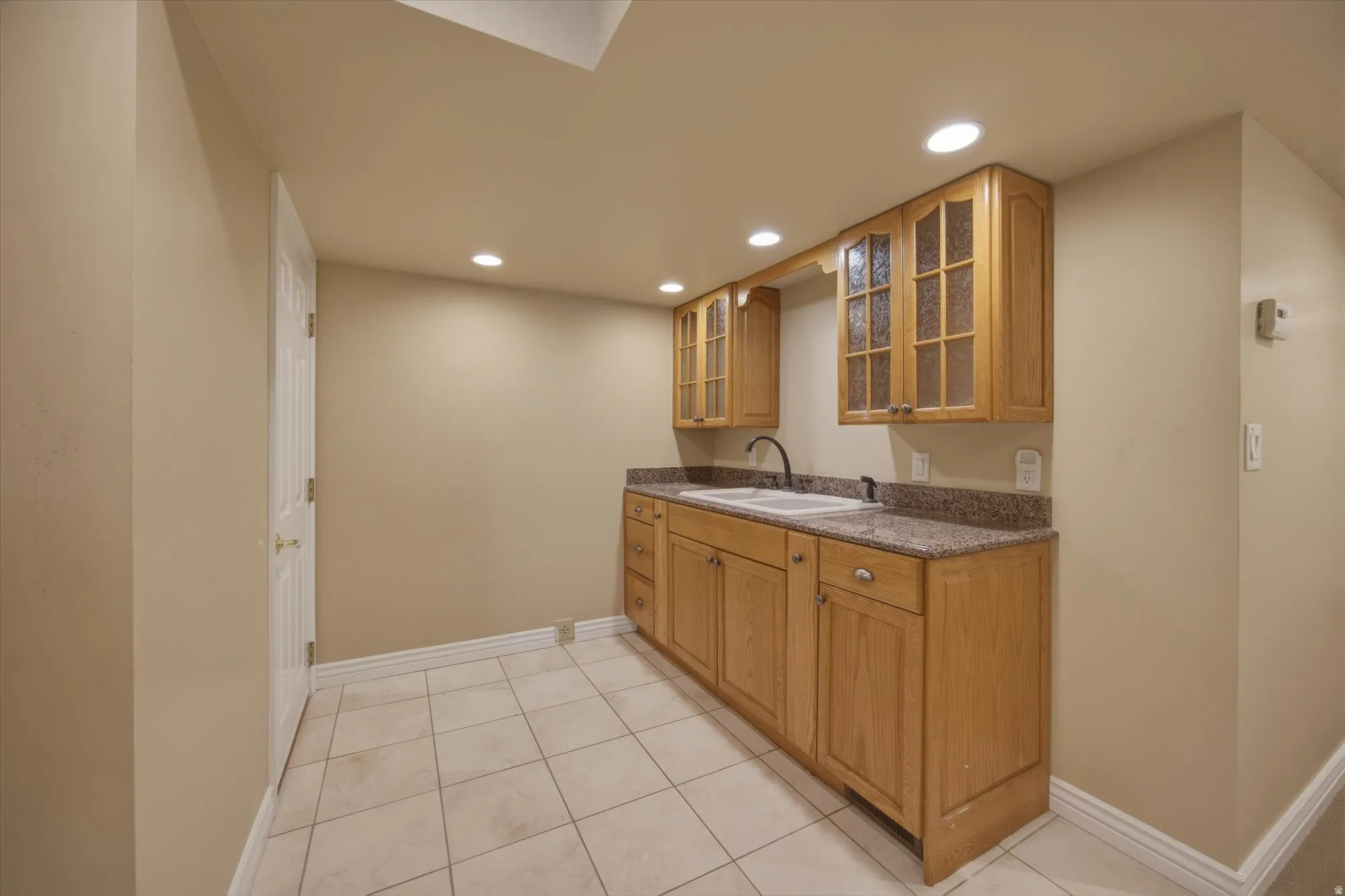 Basement kitchen featuring glass insert cabinets, recessed lighting, wood finish cabinetry, dark stone countertops, and light tile patterned floors