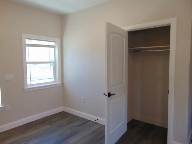 Room featuring wood-finish flooring, a double-hung window with horizontal blinds, neutral wall paint, white trim, and a bi-fold closet with a metal hanging rod and shelf