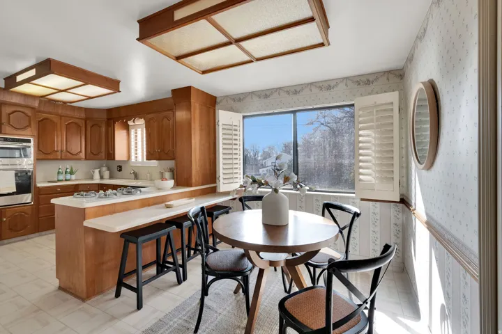 Kitchen with wallpapered walls, wood finish cabinetry, light countertops, light flooring, and a peninsula