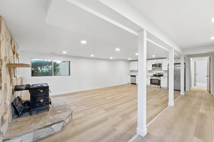Open-concept living area featuring light wood-finish flooring, recessed lighting, a stone-clad fireplace with a wood-burning stove, and a full kitchen with white cabinetry and stainless steel appliances