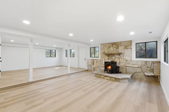 Spacious lower-level area featuring light wood-finish flooring, recessed lighting, and a stone-clad fireplace with a hearth