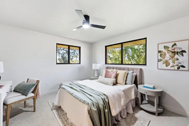 Spacious interior featuring a modern ceiling fan with integrated lighting, two large windows with black framing, light-toned carpeting, and crisp white walls