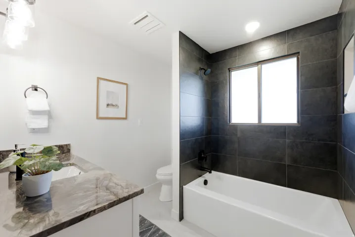 Contemporary bathroom featuring a white soaking tub with dark tile surround, a single vanity with a stone countertop, and a built-in toilet