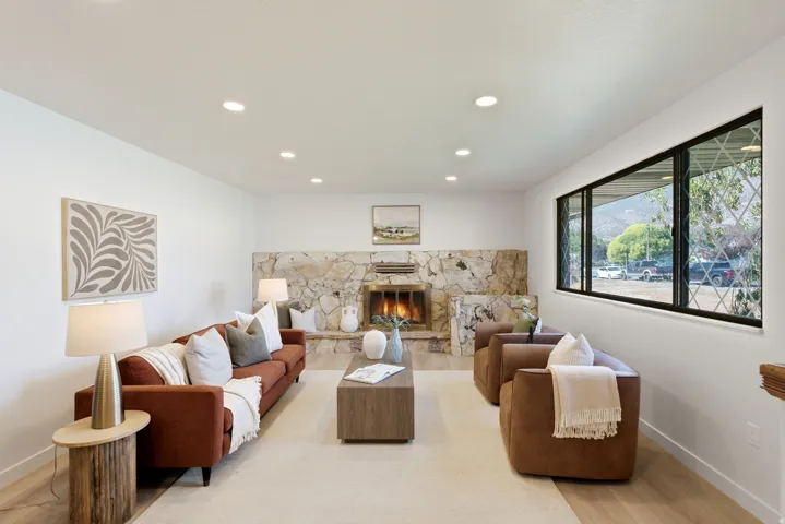 Spacious living area featuring a full-height stone fireplace, light wood-finish flooring, recessed lighting, and a large picture window with diamond-patterned security bars