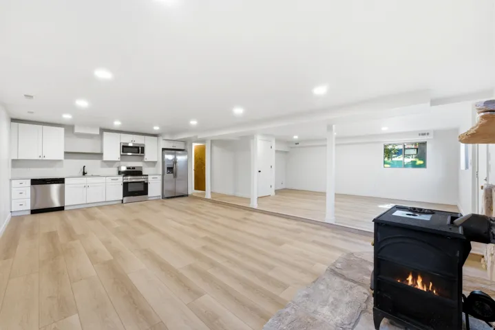 Open-concept living area featuring wood-finish flooring, a full kitchen with stainless steel appliances, recessed lighting, and a cast iron stove with a stone hearth