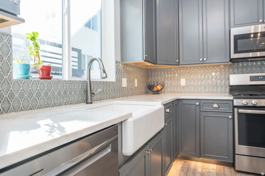 Kitchen featuring stainless steel appliances, light stone counters, decorative backsplash, light wood-style flooring, and gray cabinetry