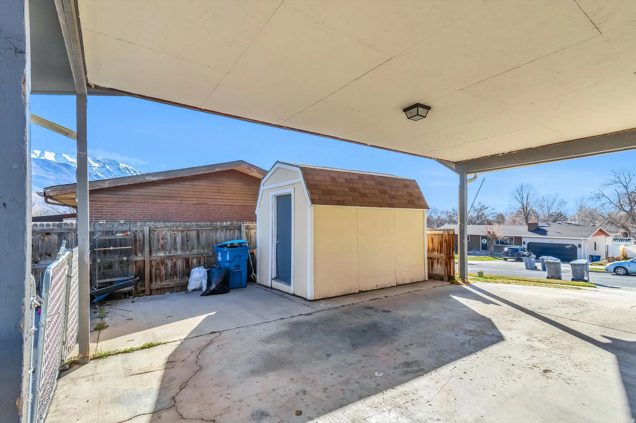 Covered carport with concrete flooring, featuring an attached exterior light fixture and a freestanding storage shed with shingle roofing
