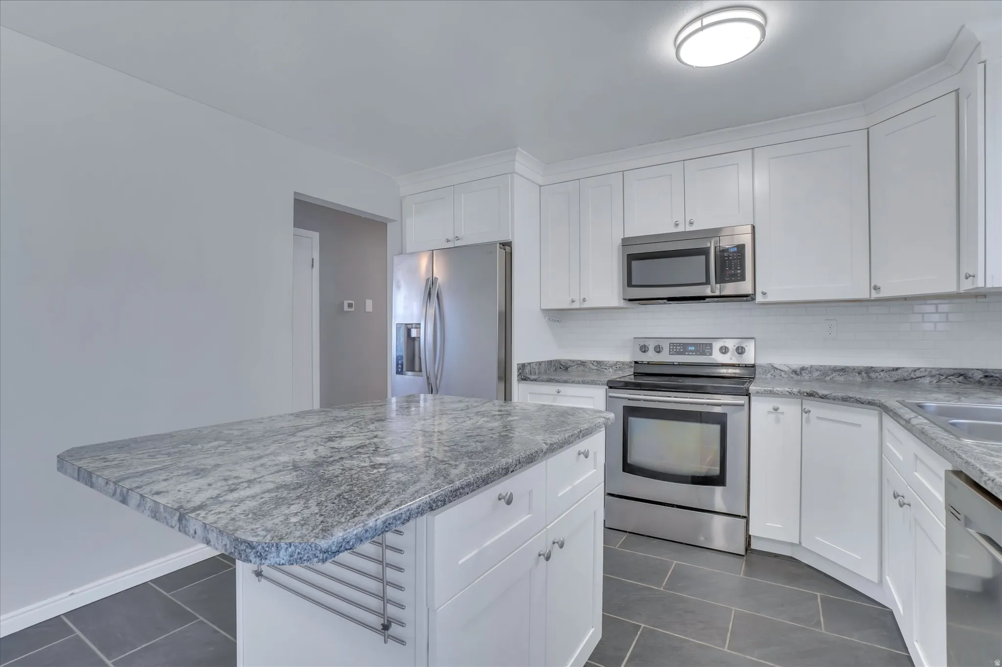 Modern kitchen featuring an island with a light-toned, patterned countertop, white cabinetry, stainless steel appliances, a white subway tile backsplash, and dark rectangular floor tiles