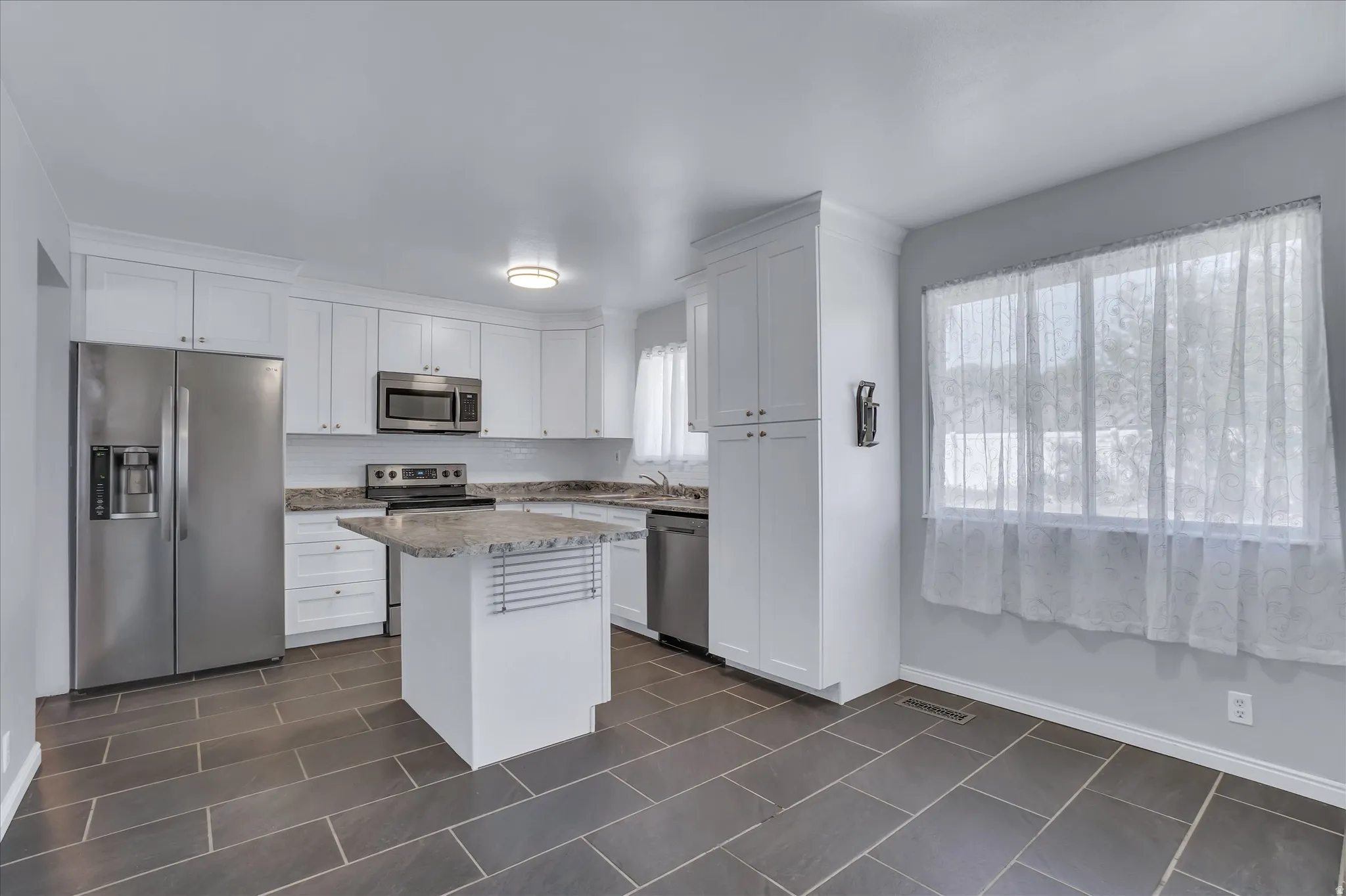 Modern kitchen featuring white cabinetry, stainless steel appliances, a center island with a granite-style countertop, a tile backsplash, and dark tile flooring