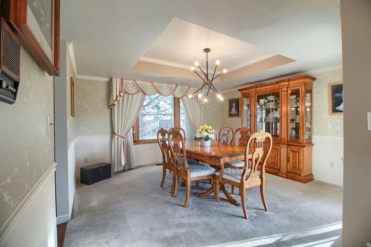 Dining area featuring wallpapered walls, a raised ceiling, ornamental molding, a chandelier, and light colored carpet