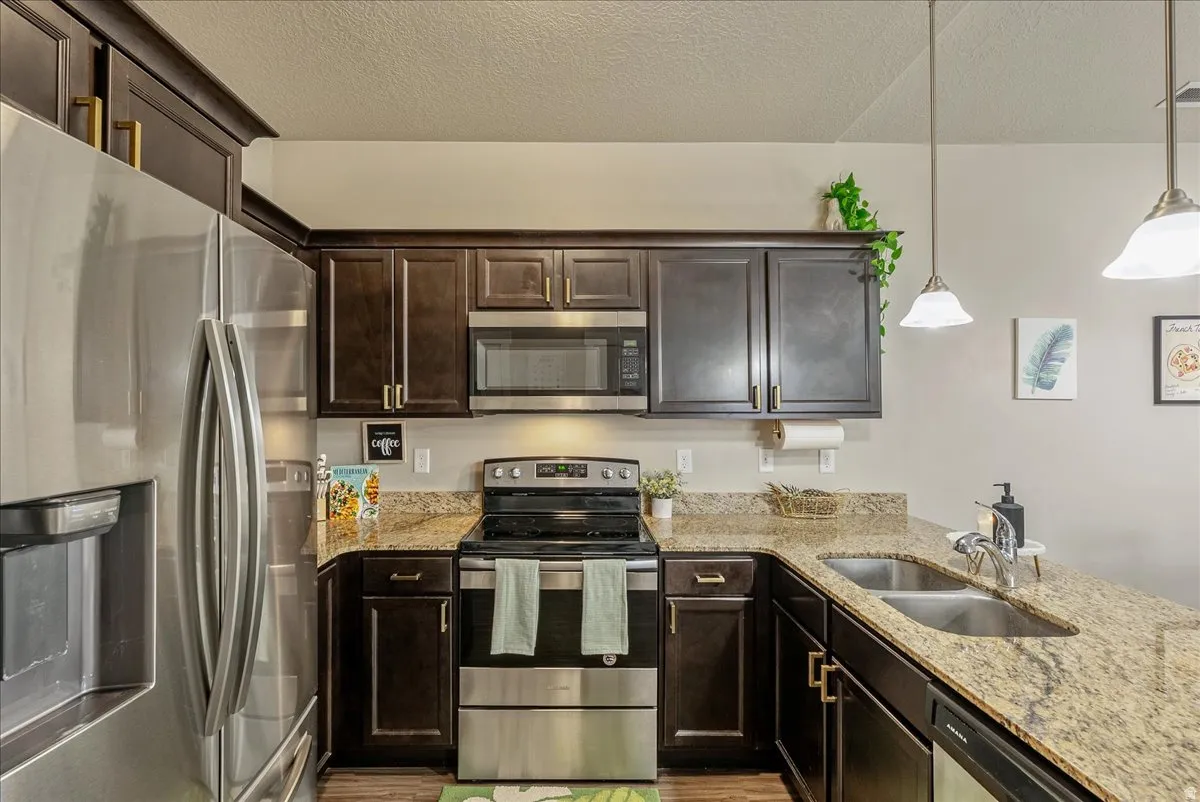 Kitchen featuring stainless steel appliances, dark wood finish cabinetry, light stone counters, hanging light fixtures, and a textured ceiling
