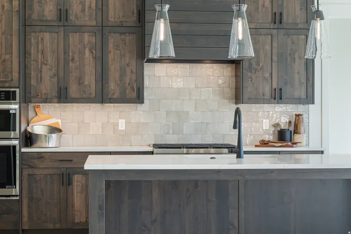 Kitchen featuring dark wood finish cabinetry, backsplash, stainless steel double oven, light stone countertops, and pendant lighting