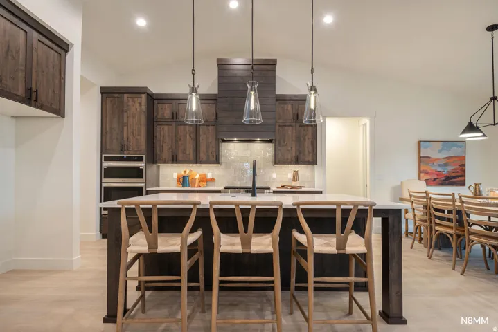Kitchen featuring dark wood finish cabinets, stainless steel double oven, an island with sink, light stone countertops, and a breakfast bar